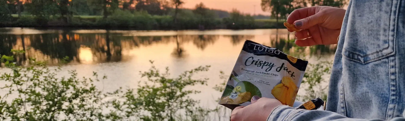 In der Kategorie Exotische Fruchtchips präsentiert Lotao Jackfruit Chips, hier im Kategoriebanner Desktop mit einem Model, das vor stillem Gewässer im Sonnenuntergang in eine Tüte mit Chips greift.