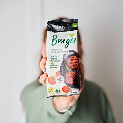 Hand hält eine 100g Packung vom Lotao Veggie Burger Mix, dem veganen Fleischersatz für saftige vegetarische Burger auf Basis von Jackfruit und Erbsen in Bio-Qualität. 