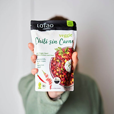 Hand hält 320g Packung der Lotao Veggie Chili sin Carne Bowl, einem bio-veganen Fertiggericht mit Lotao Veggie Hack, Kidneybohnen und Mais als vegane Bio-Mahlzeit