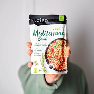 Hand hält 220g Packung der Lotao Veggie Mediterranen Bowl, einem bio-veganen Fertiggericht mit Buchweizen und getrockneten als vegane Bio-Mahlzeit
