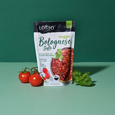 320g Packung Fertigsauce Veggie Bolognese Sosse von Lotao mit bio-veganer Fertigsosse Bolognese mit Lotao Veggie Hack als veganer Fleischersatz und Tomaten vor grünem Hintergrund